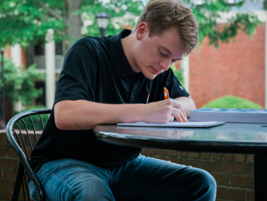 Student writing on a notepad while sitting at a table outside